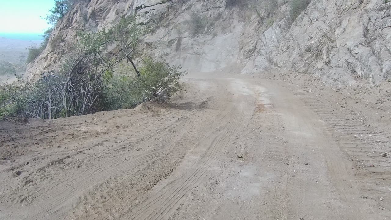 The road bends through dry mountain terrain in Los Cabos, with a dusty track, exposed rock, low brush near the edge, and no visible people.