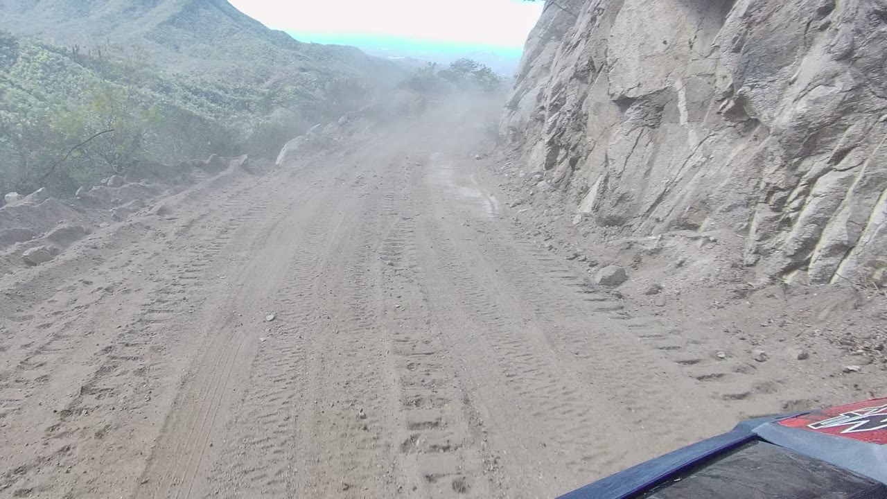 Opening view of a dusty unpaved road in Los Cabos, Baja California Sur, with tire tracks in the foreground, a pale rock face to the right, green hills ahead, and no visible people.