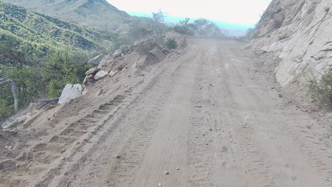 The road in Los Cabos runs past a rocky cut with scattered stones on the dirt surface, scrub and green hills visible beyond the edge, and no visible people.