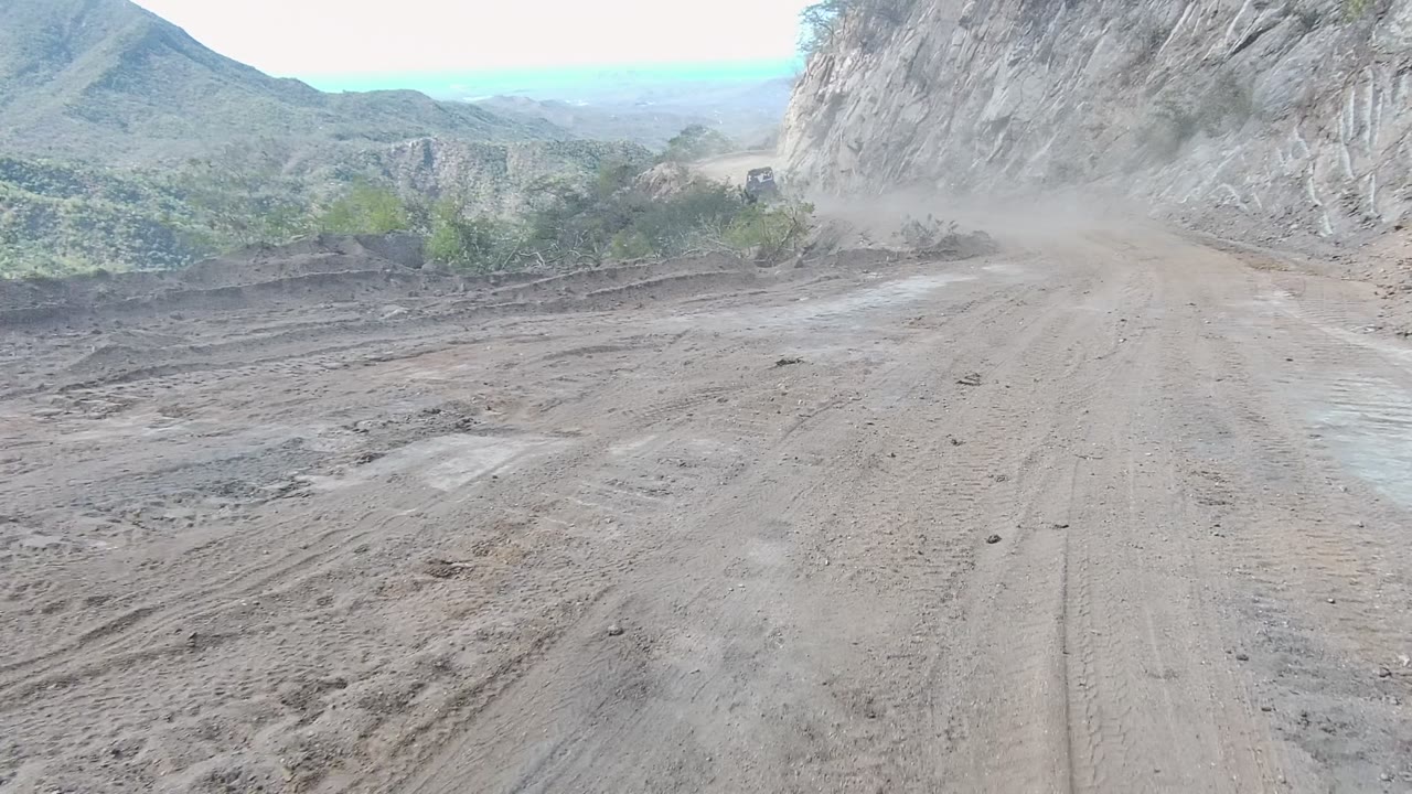 Forward view of a dusty mountain dirt road in Los Cabos, Baja California Sur, with tire tracks, rocky slopes, distant hills, scrub vegetation, and no visible people.