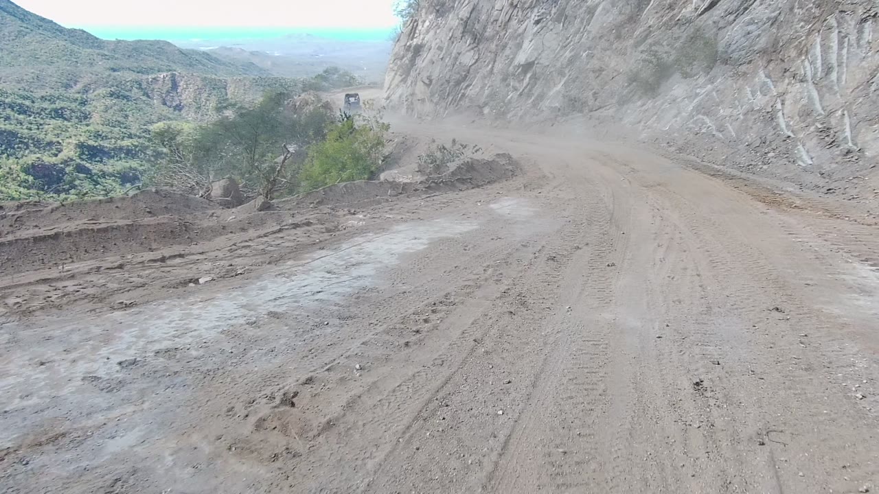 Dirt road narrowing around a rock wall with dust ahead.