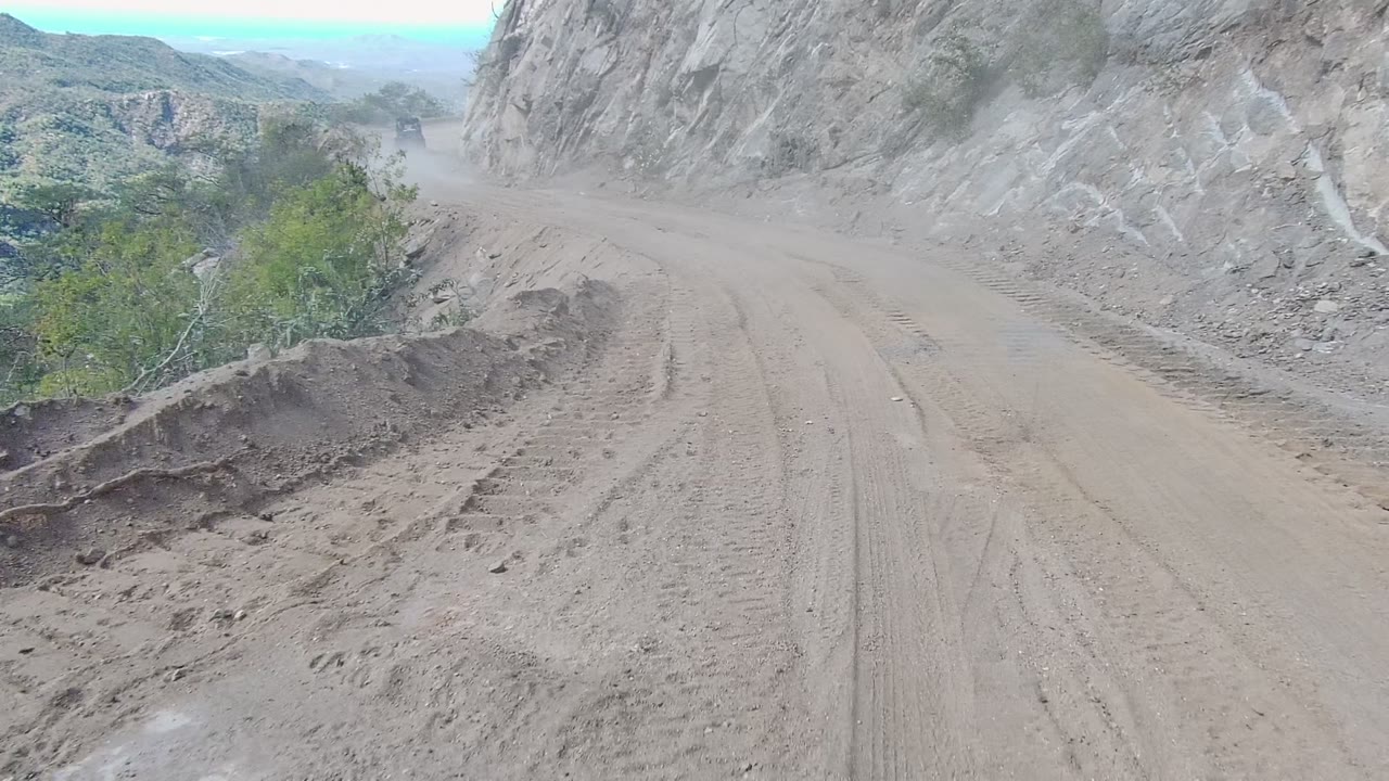 A forward vehicle perspective on a dusty Los Cabos dirt road shows rutted tire marks, a gray rock face to the right, low green shrubs, hazy hills, and no visible people.