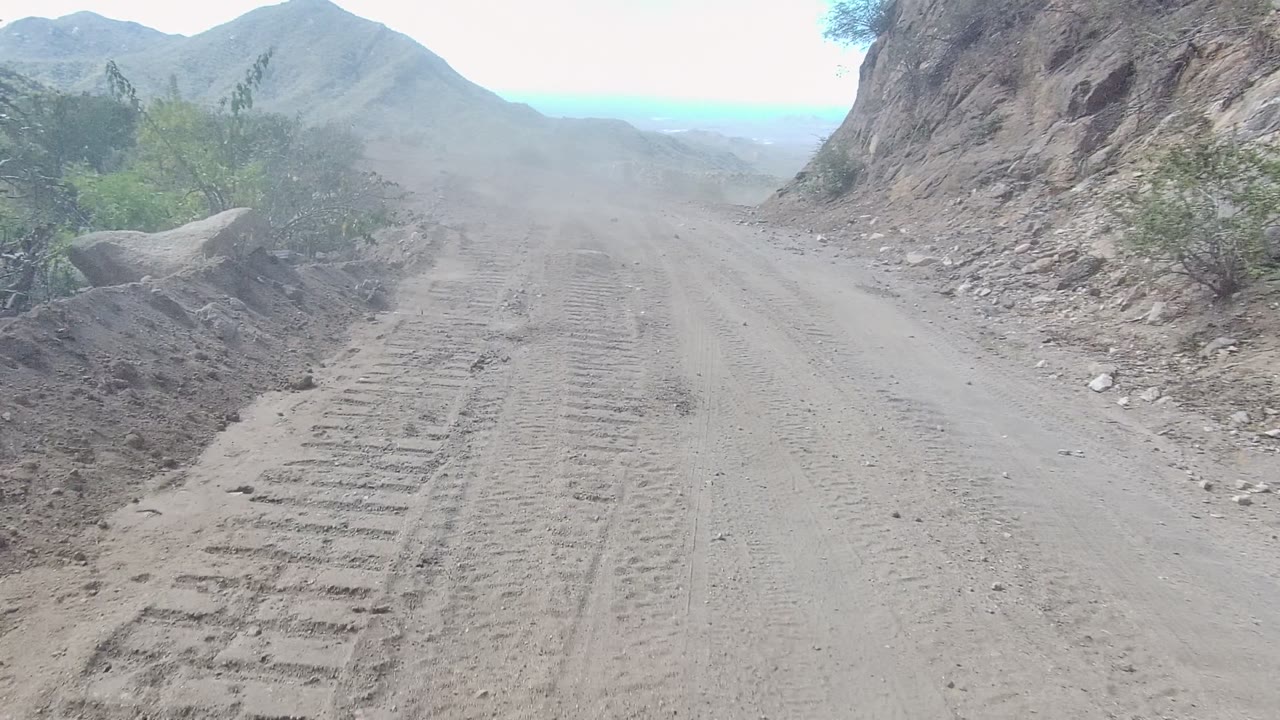 Los Cabos dirt road seen from vehicle height, with tire tracks in the foreground, rocky slopes on both sides, distant hills, pale sky, and no visible people.