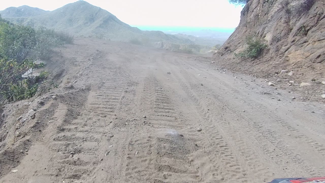 Vehicle-height view along a rutted dirt road in Los Cabos, with a rock face on the right, scrub along the edges, layered hills ahead, and no visible people.