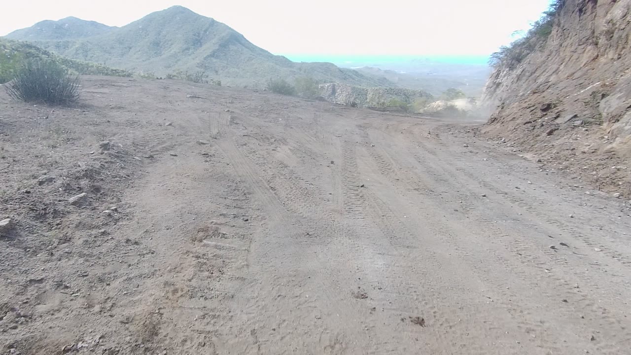 Los Cabos mountain dirt road opening into a wider bend, with tire marks across the dusty surface, a rock wall to the right, distant hills, a small dust plume, and no visible people.