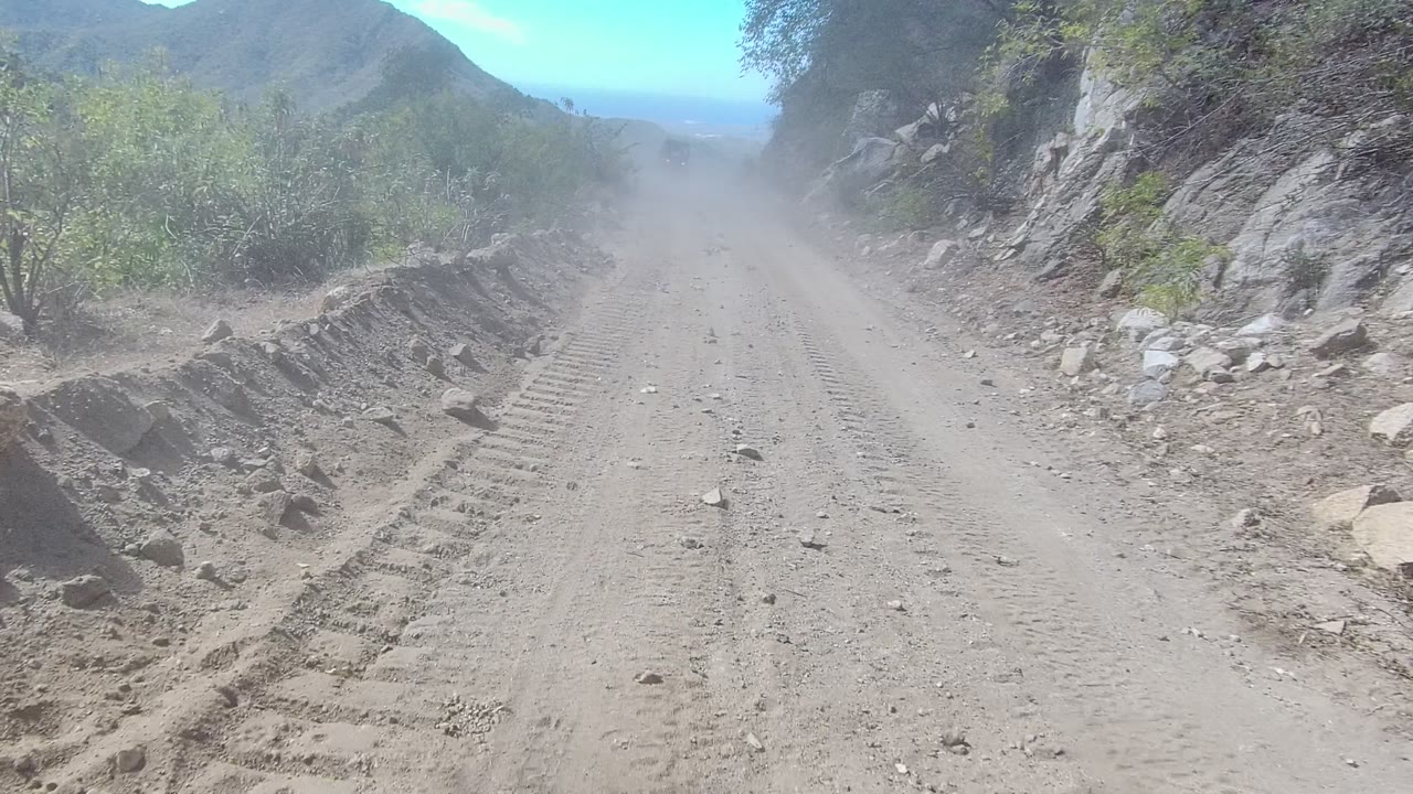 The road continues through rocky Los Cabos mountain terrain, with compacted dirt, scattered stones, green roadside brush, hazy distance, and no visible people.