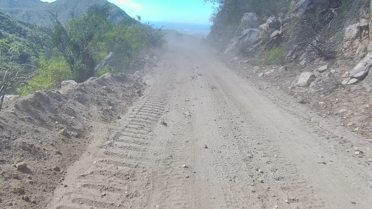 A forward view down the Los Cabos dirt road shows tire impressions leading into dust, with a rocky wall on one side, shrubs on the other, and no visible people.