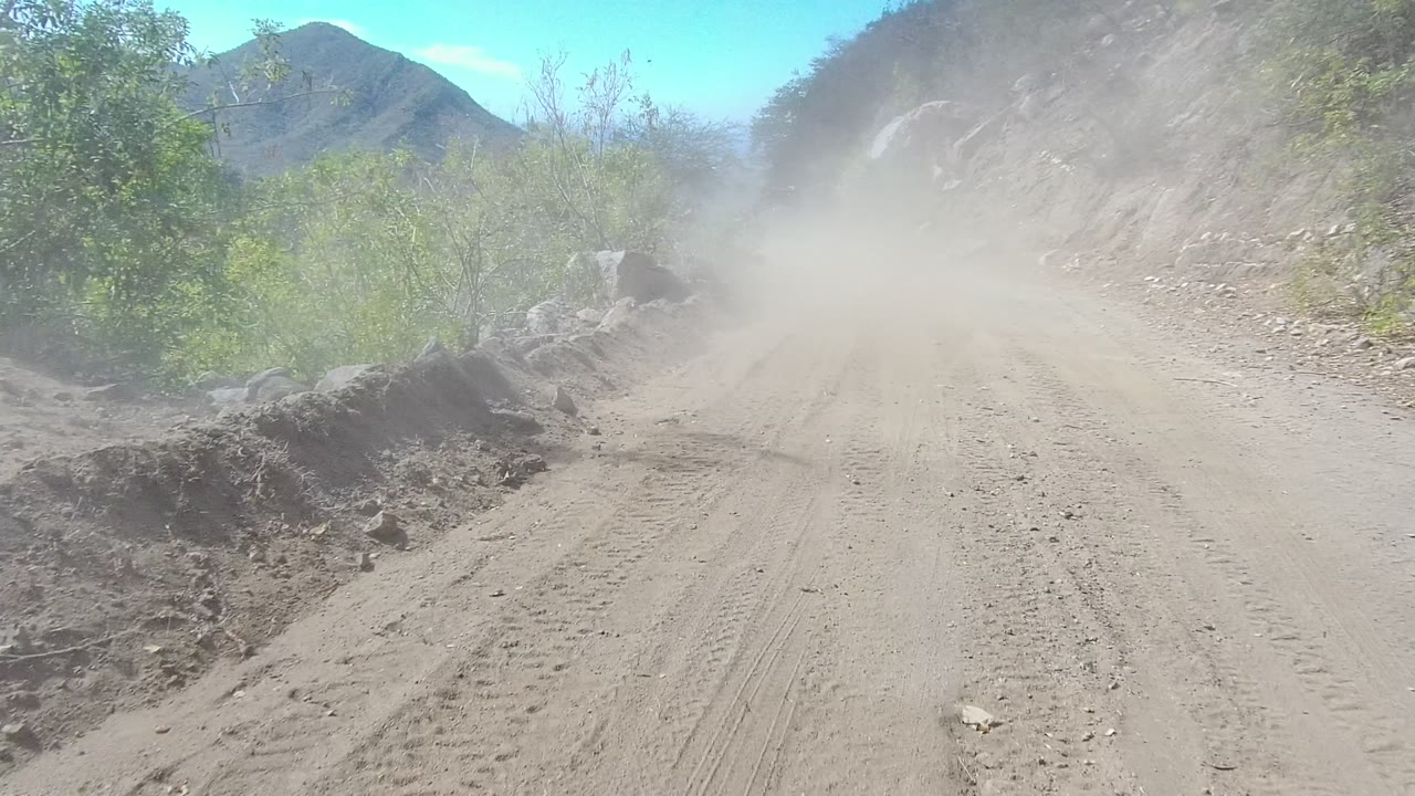A pale dirt road bends beside a rocky embankment and green roadside brush in Los Cabos, with a broad dust plume reducing background detail and no visible people.