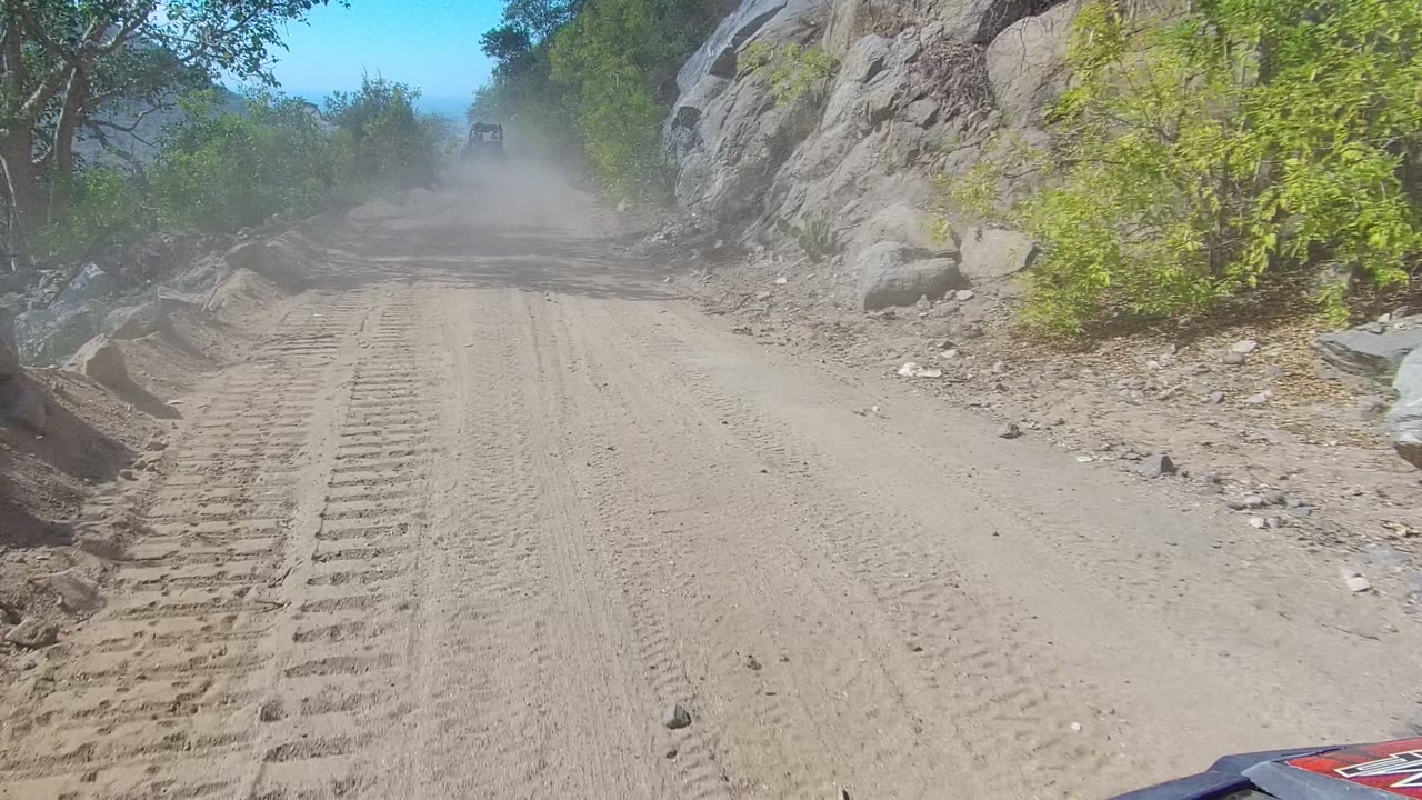 Wide dirt road in Los Cabos with heavy tire tread marks across the foreground, rocks and trees along the sides, dust ahead, and no visible people.