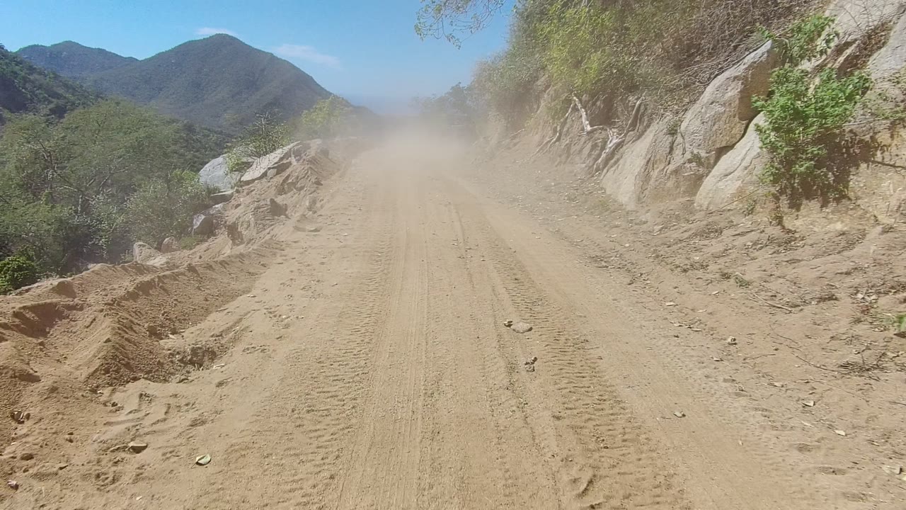 Tire-marked dirt road in Los Cabos, Baja California Sur, with dust hanging over the track, rocky slope at right, green mountains in the distance, and no visible people.