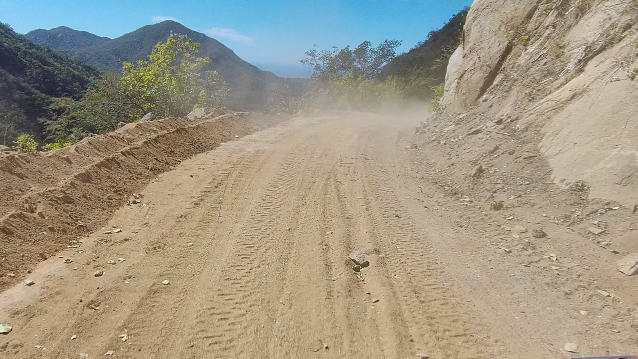 The Los Cabos dirt road passes a rough berm and exposed rock face, with tan dust drifting across the lane, scrub vegetation along the edges, and no visible people.