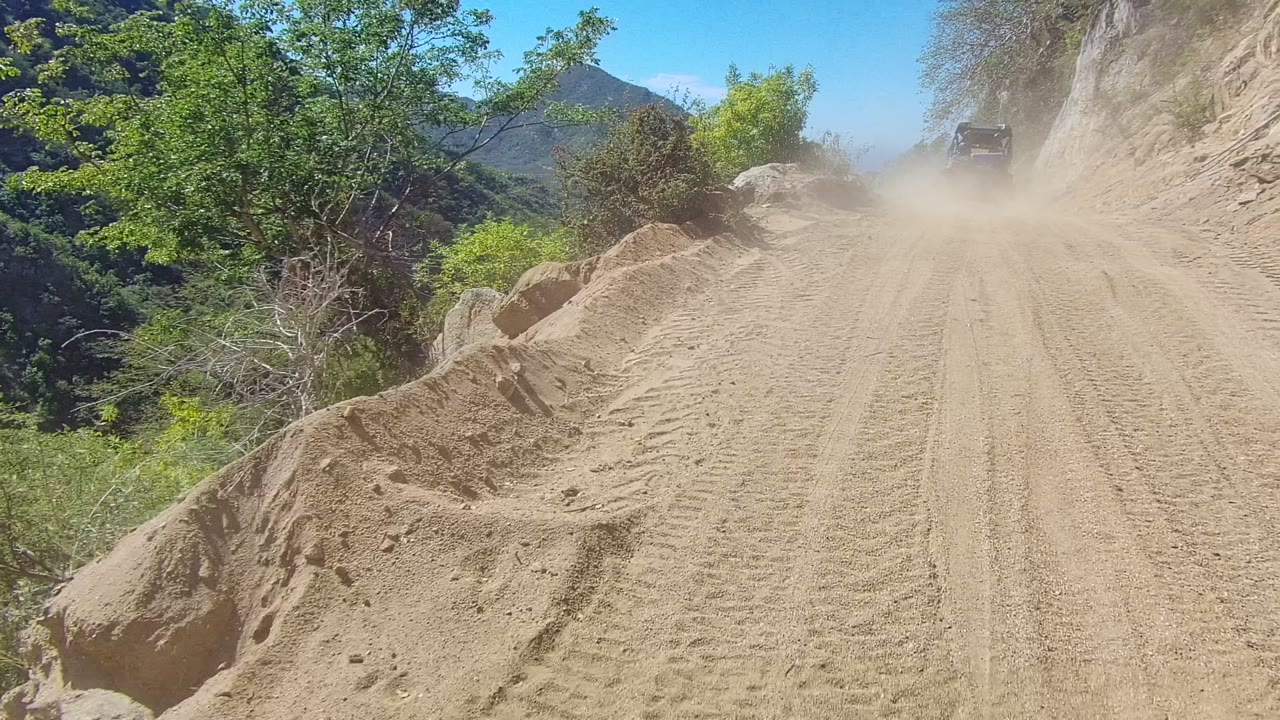 Opening frame of a sandy dirt road in Los Cabos, Baja California Sur, with tire tracks, roadside rocks, green vegetation, distant mountains, and no visible people.
