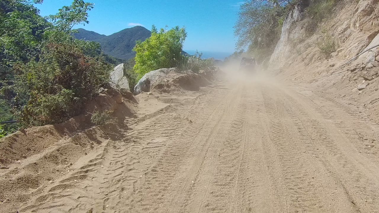 Dust cloud on a dirt mountain road beside a rocky slope.