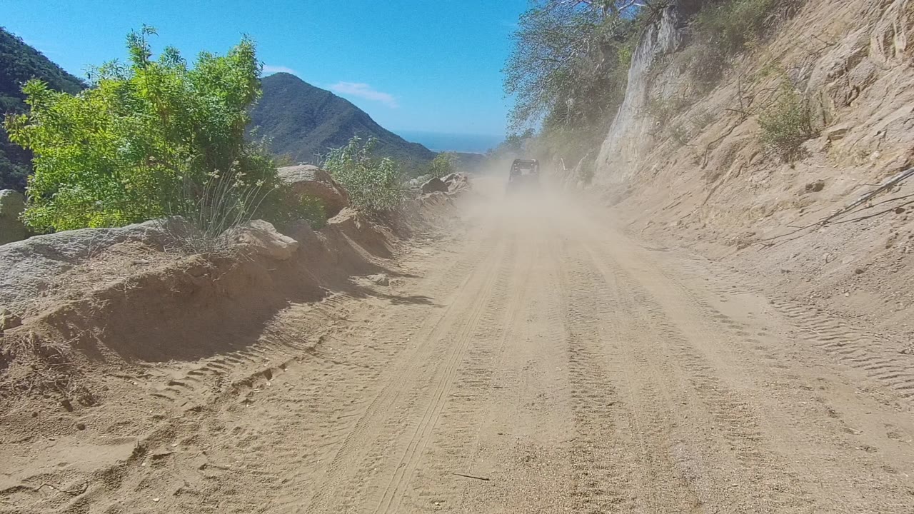 A small vehicle shape is partly visible through dust on a sandy Los Cabos mountain road, with boulders, shrubs, a steep rock wall, and no visible people.