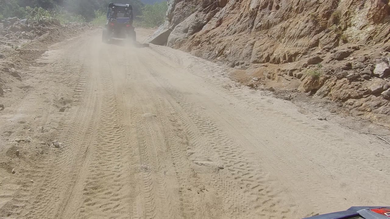 At 0:00, a dark off-road vehicle drives away on a dusty sandy road in Los Cabos, Mexico, with tan rock walls, tire tracks, and no visible people.