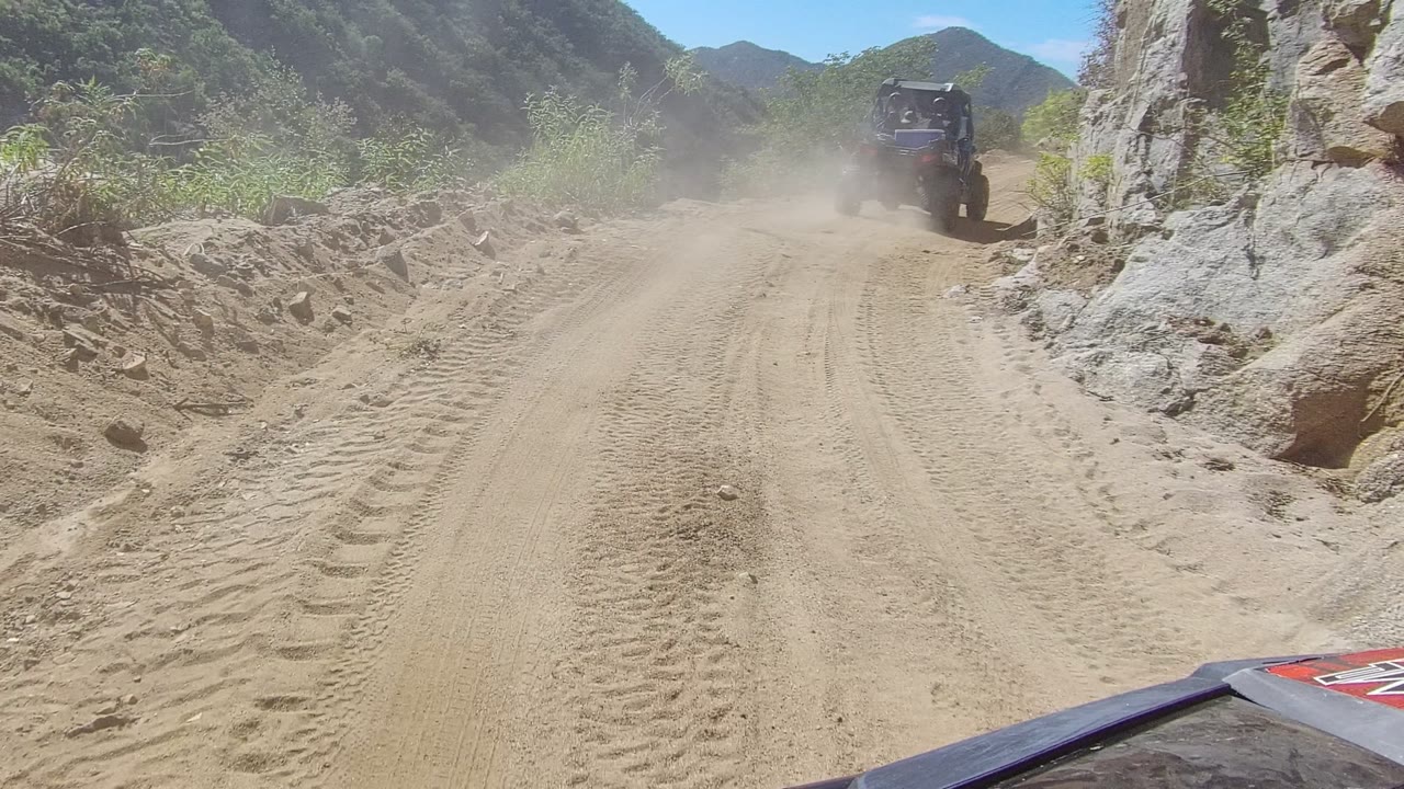 At 0:02, the road opens slightly between rocky banks and green mountain slopes in Los Cabos, Mexico, as the off-road vehicle moves ahead with no visible people.