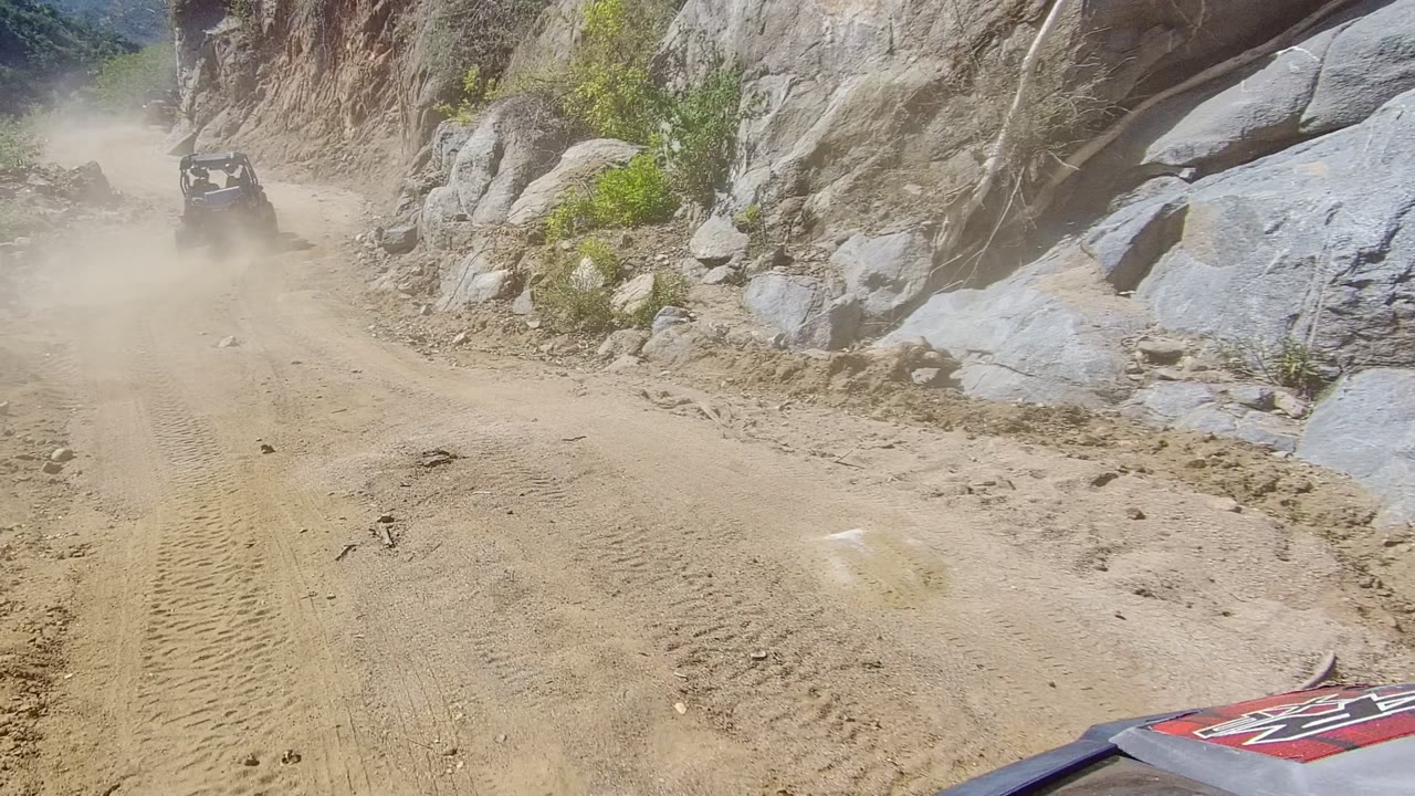 An off-road vehicle appears ahead on the Los Cabos mountain trail, kicking up dust between rocky walls with no visible people.