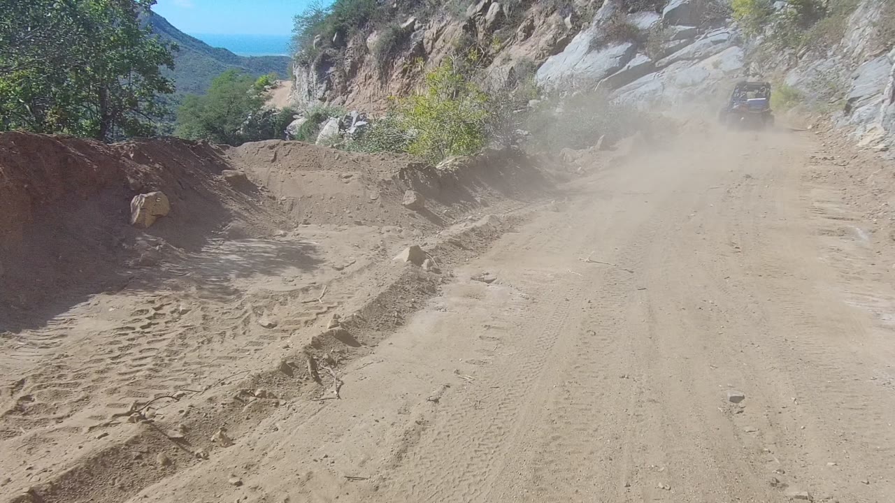 Sandy mountain road with tire tracks, rocks, shrubs, and dust from a vehicle ahead.