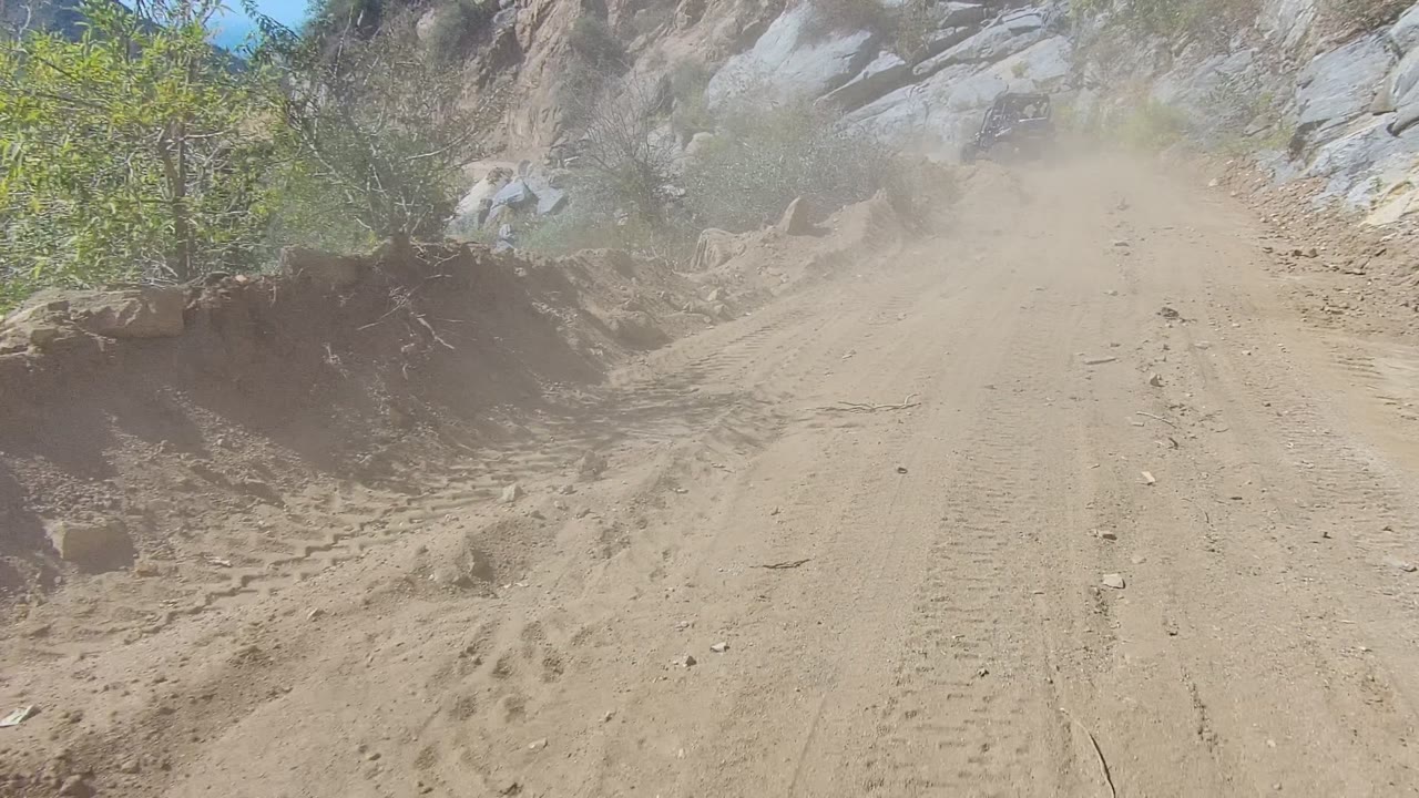 The Los Cabos trail narrows between pale rock faces and scrub, with a vehicle barely visible through tan dust and no visible people.