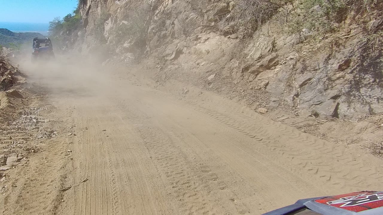 Los Cabos dirt road viewed from a following vehicle, with a dust cloud around a vehicle ahead, rocky cliff walls on the right, distant mountains, and no visible people.