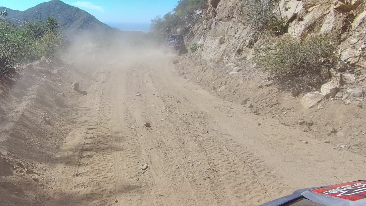 Los Cabos dirt track between rocky slopes, with a hazy dust cloud softening the road ahead, sparse shrubs at the edges, distant mountain ridges, and no visible people.