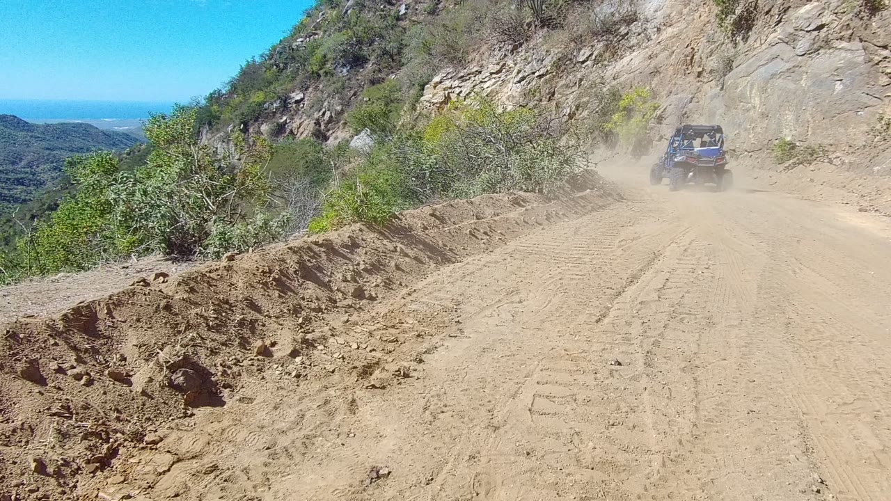 A dark 4x4 drives away on a Los Cabos mountain dirt road, with a dust trail, scrub on the left, rocky hillside on the right, distant blue horizon, and no visible people.