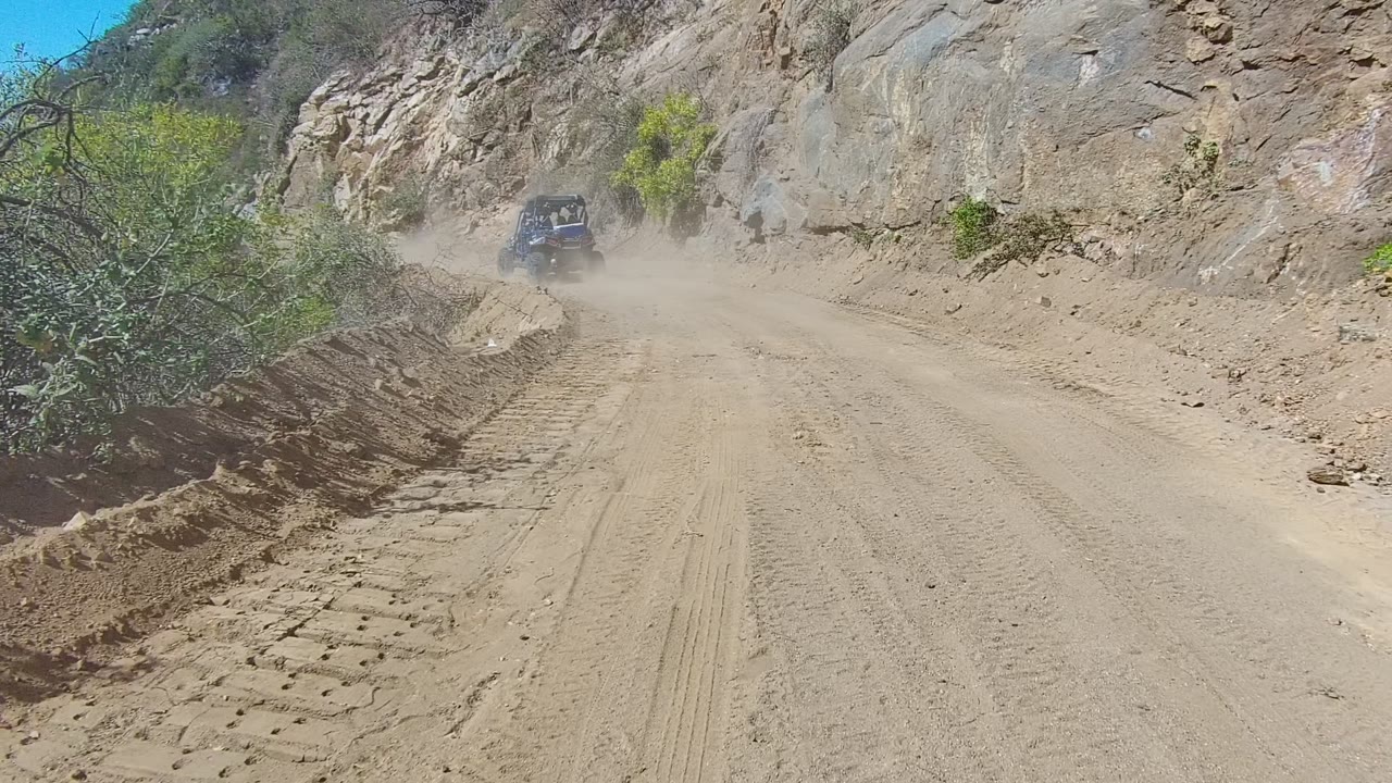 A dark 4x4 appears farther ahead on the Los Cabos dirt road, surrounded by airborne dust, with a rocky cut slope on the right, brush on the left, and no visible people.