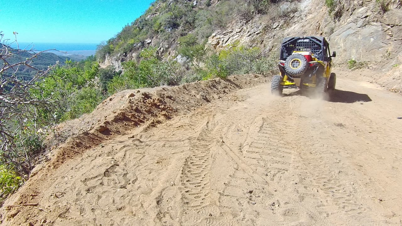 At 0:00 in Los Cabos, a yellow and black off-road vehicle drives away on a sandy mountain road beside shrubs and rocky hillside, with no visible people.