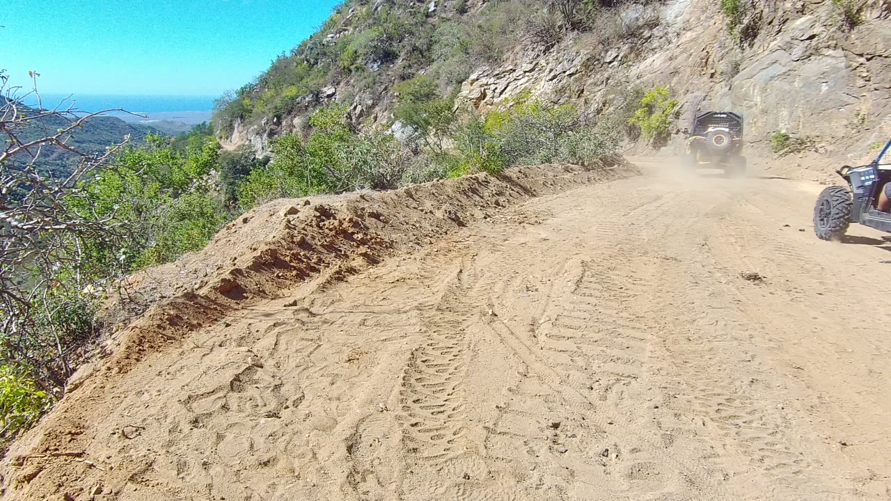 At 0:02 in Los Cabos, dust hangs over the mountain road as the first vehicle exits near the right edge and the sandy track remains open in foreground, with no visible people.