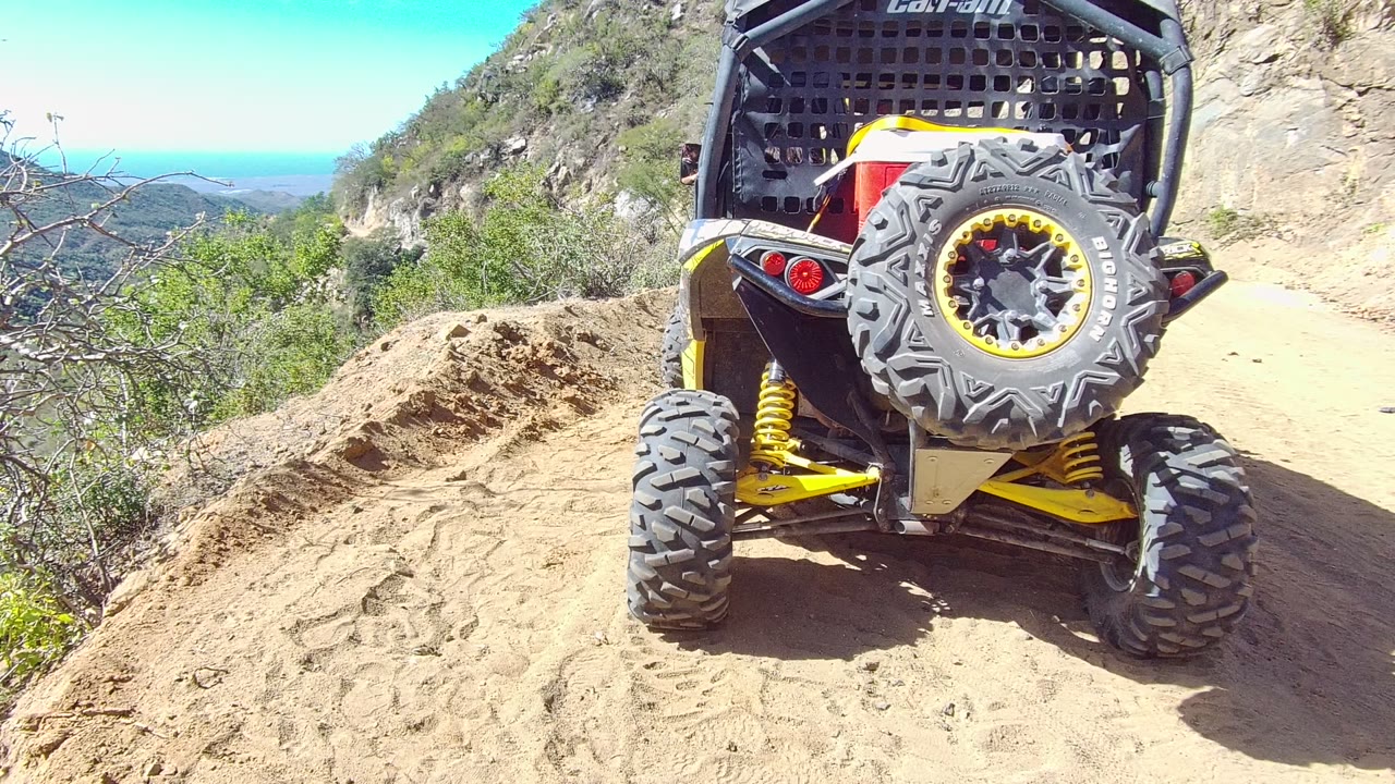 Rear close view of a yellow and black UTV starting up a sandy mountain trail in Los Cabos, Baja California Sur, with rocky slope, scrub vegetation, blue sky, distant water, and no visible people.