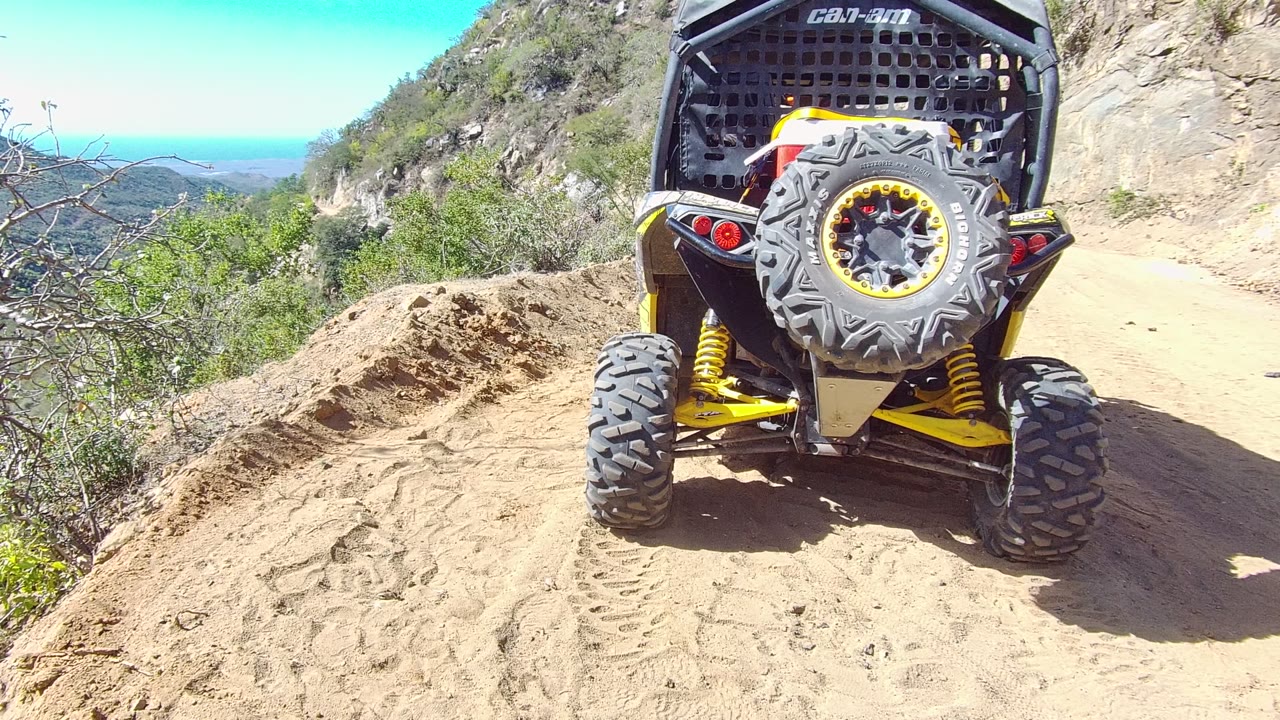 Rear view of the UTV moving farther up the narrow Los Cabos dirt trail, with sandy berms, scrub brush, a rocky hillside, blue sky, and no visible people.
