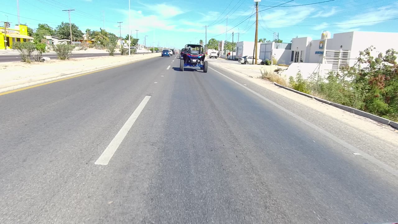 Forward road view in Los Cabos, Baja California Sur, with a dark off-road vehicle ahead, low roadside buildings, parked cars, utility poles, and no visible people.