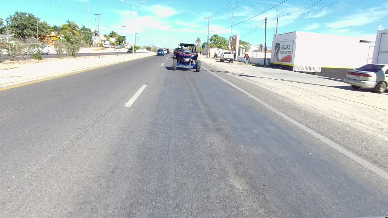 Forward-facing Los Cabos street view with an off-road vehicle ahead, a roadside business wall on the right, a parked car, utility poles, and no visible people.