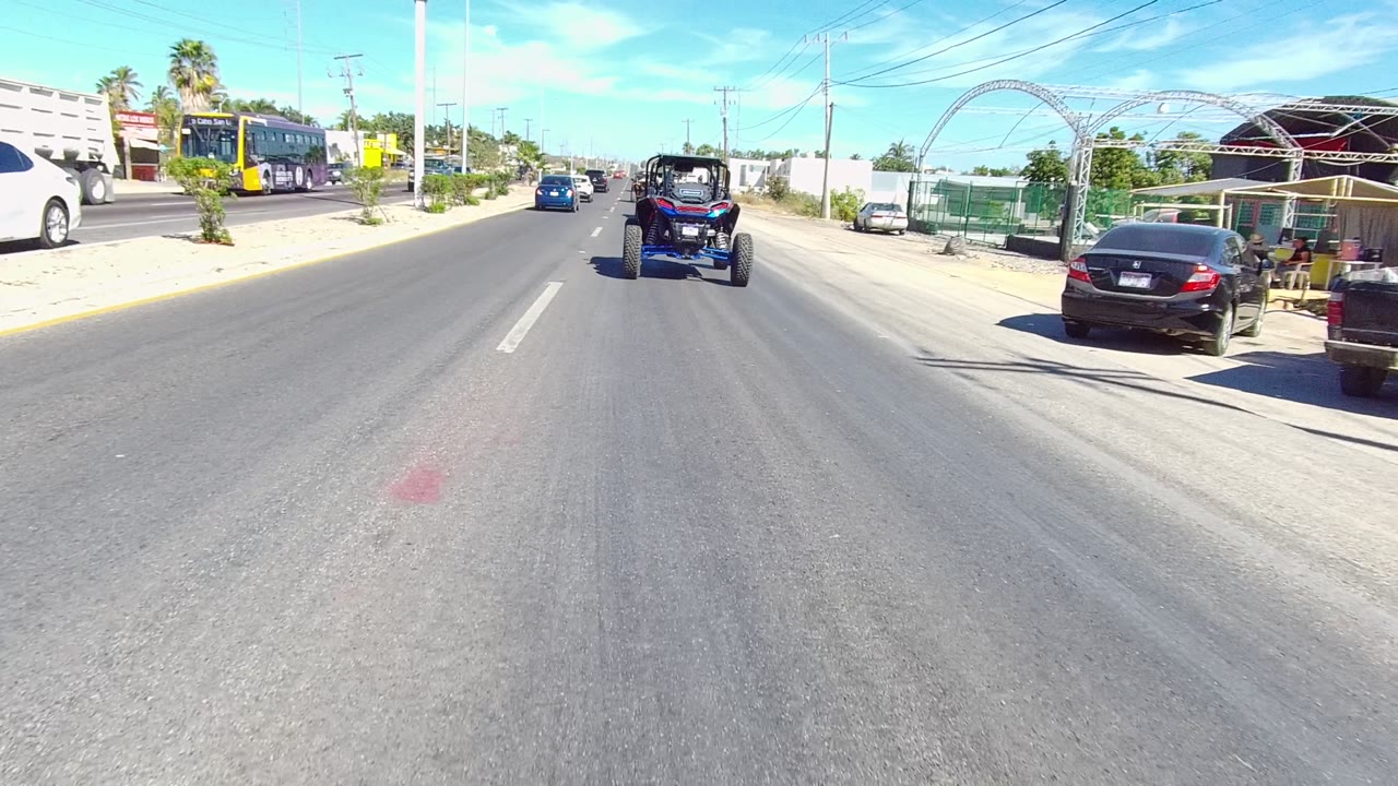 A dune buggy drives ahead on a paved road in Los Cabos with a parked black car, a bus, palm trees, low buildings, and no visible people.