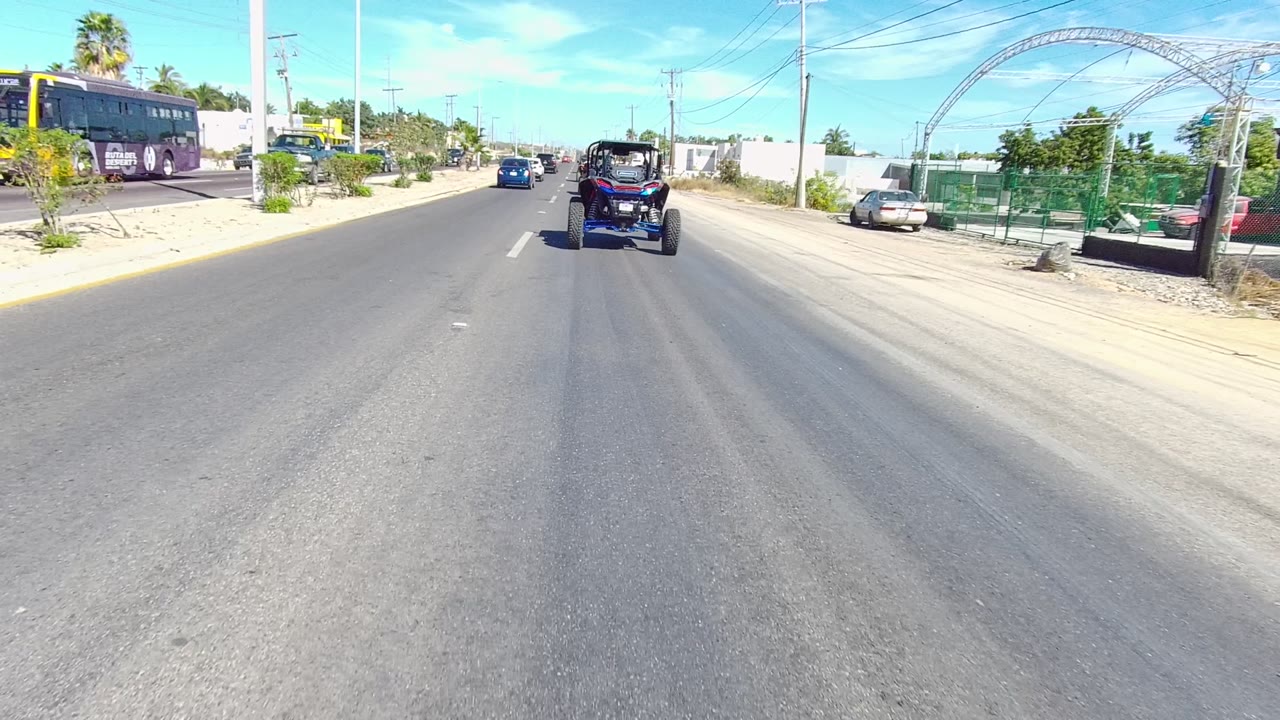 Dune buggy centered on a sunlit road with a bus and arched roadside structure.