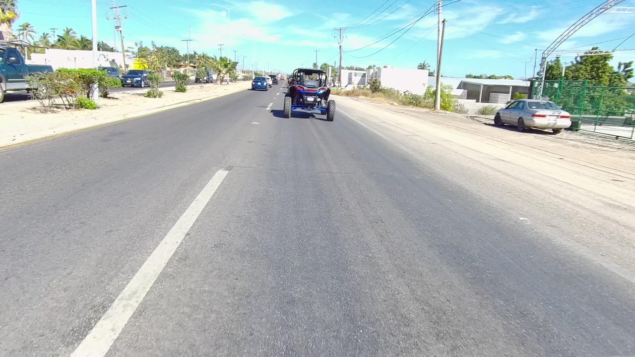 A dune buggy travels farther down the paved road in Los Cabos, framed by white roadside walls, parked cars, palm trees, overhead wires, and no visible people.