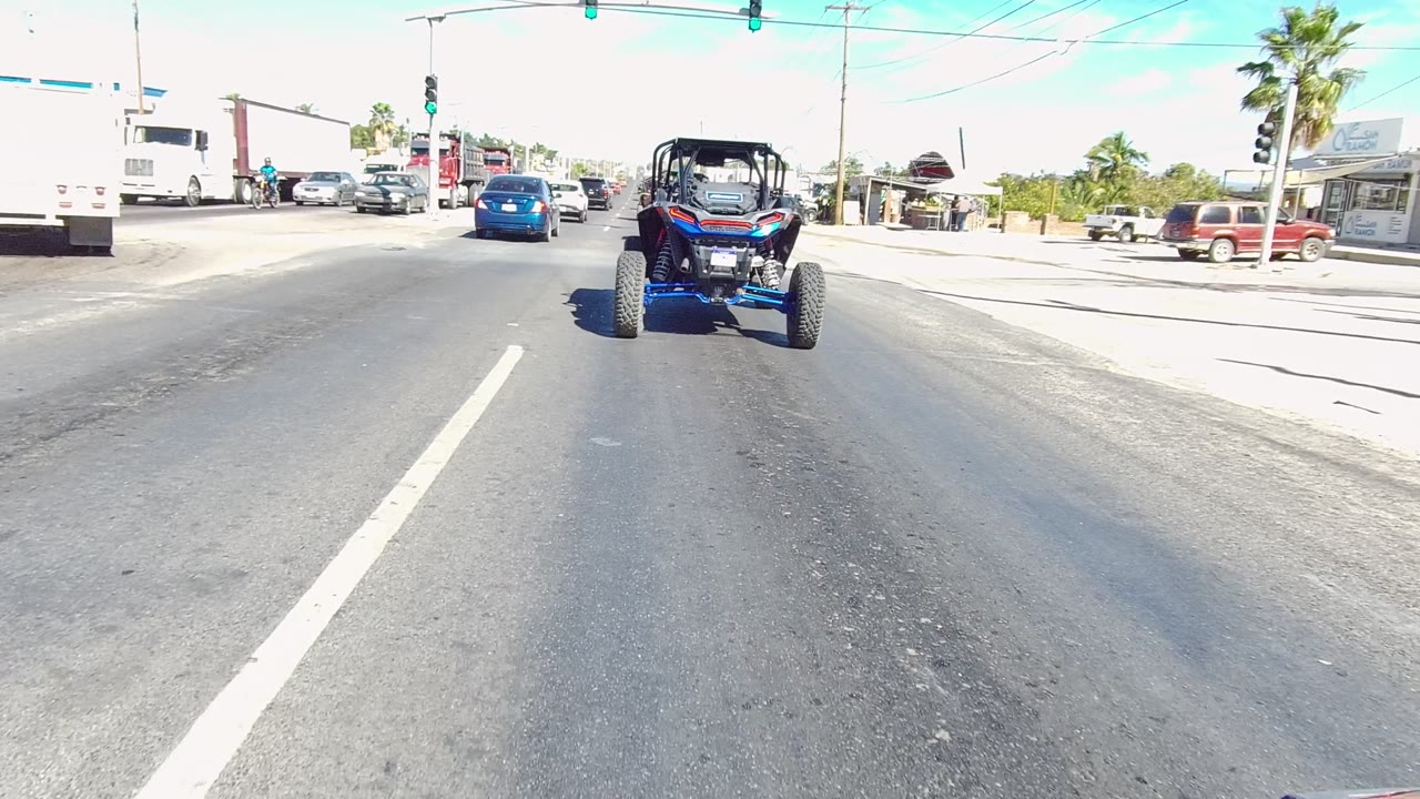A blue UTV drives ahead on a Los Cabos road beneath traffic lights, with cars and trucks nearby and no visible people in this opening frame.