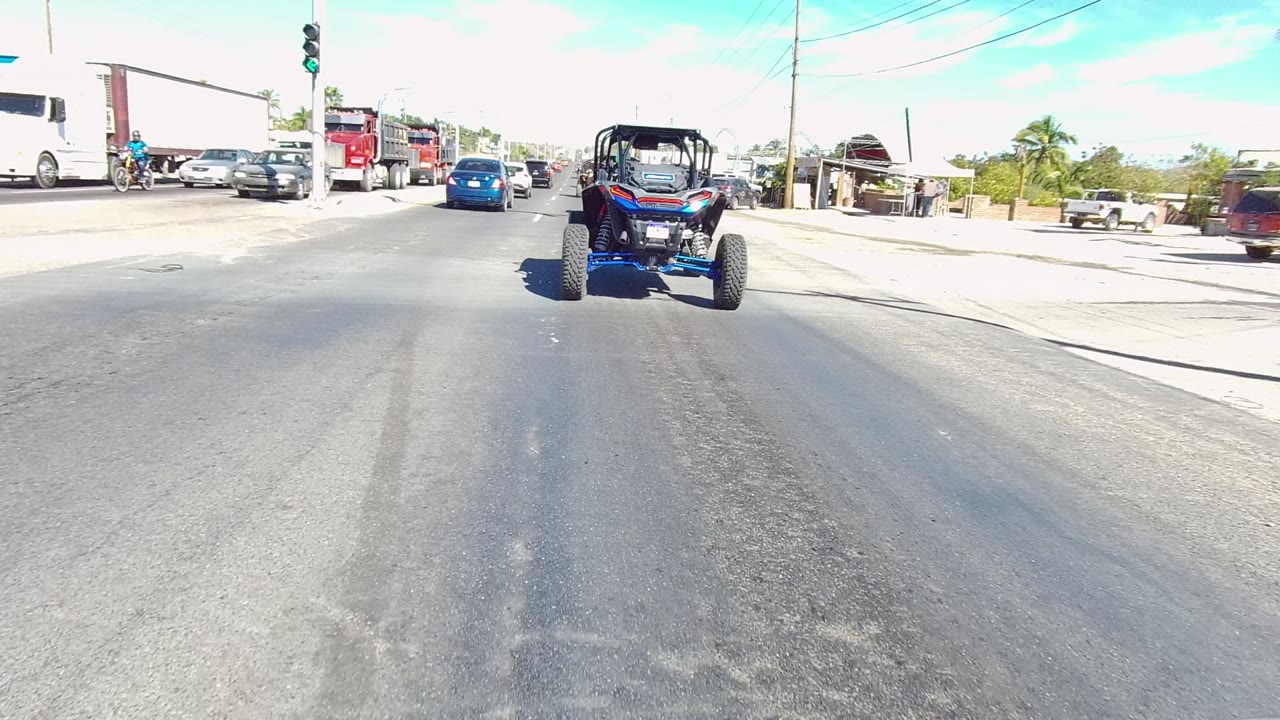 Blue UTV ahead with large trucks and cars on a sunny street