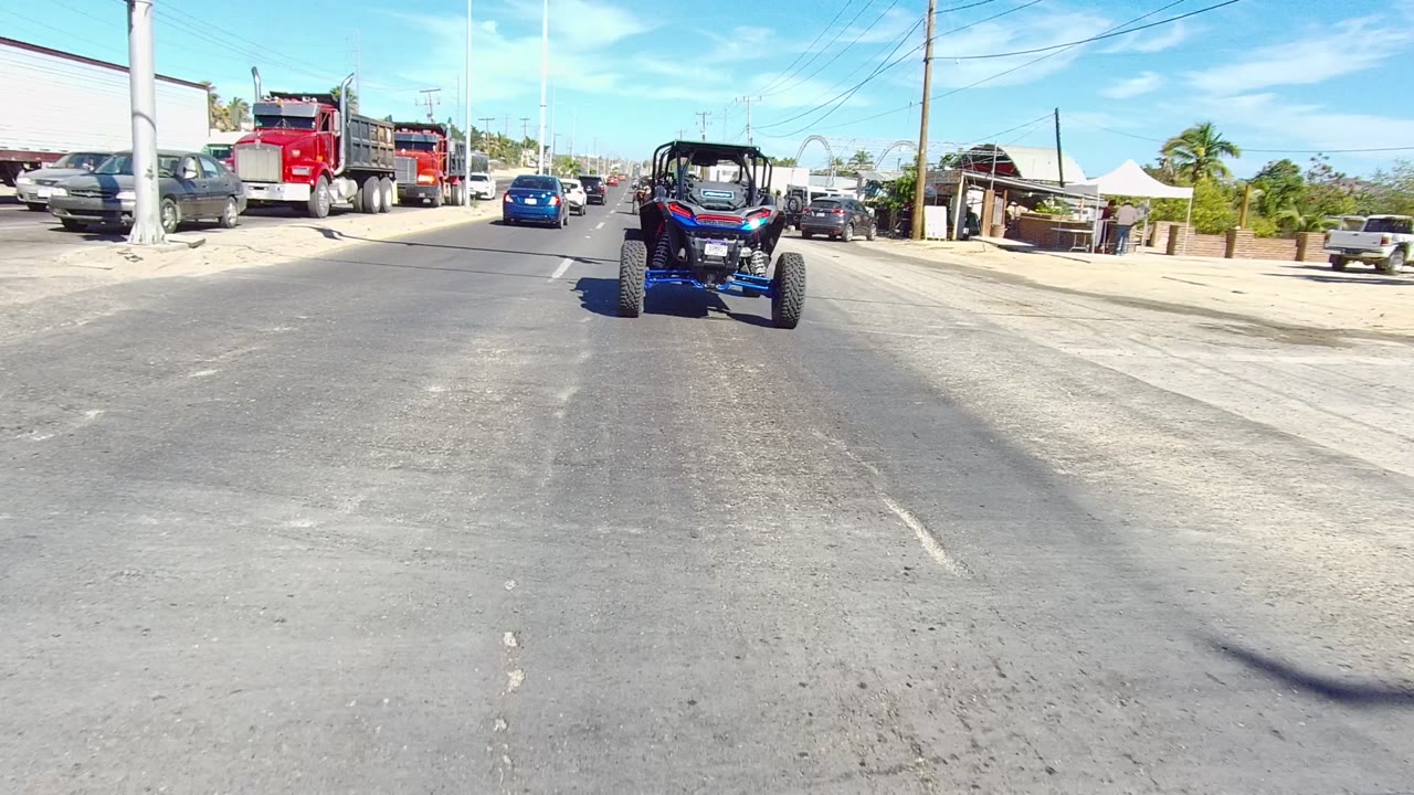 A blue UTV travels ahead on a Los Cabos street while red dump trucks, cars, lane markings, overhead signals, and palm trees fill the surrounding traffic scene, with no visible people.
