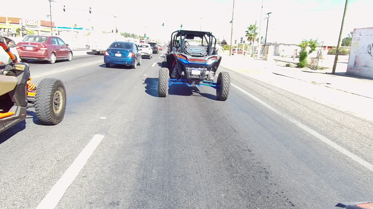 Blue off-road vehicle centered ahead on a Los Cabos road, with a blue sedan, red car, white van, traffic lights, and no visible people.