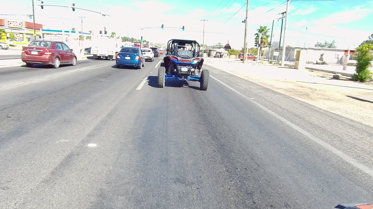 A wider moment on the Los Cabos roadway shows the off-road vehicle slightly right of center, the red car to the left, a blue sedan ahead, and no visible people.