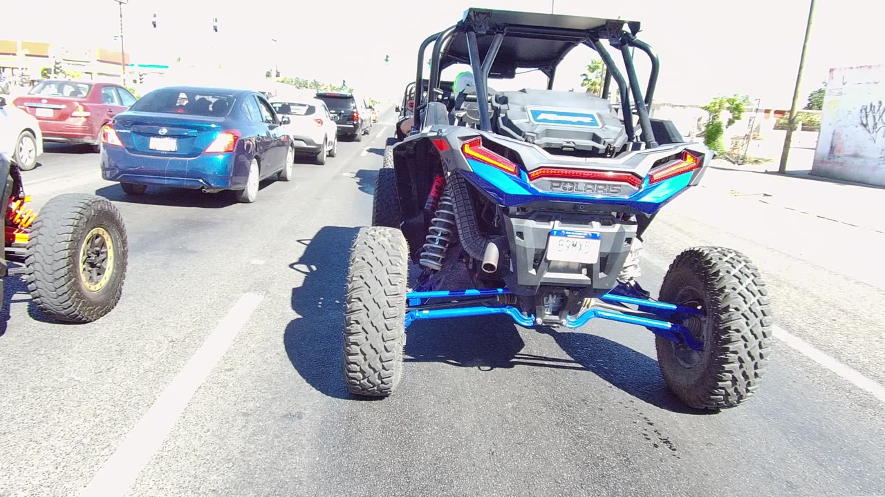 At 0:02 in Los Cabos, the rear of the UTV dominates the right side of the frame while cars occupy the left and center lanes under bright daylight, with no visible people.