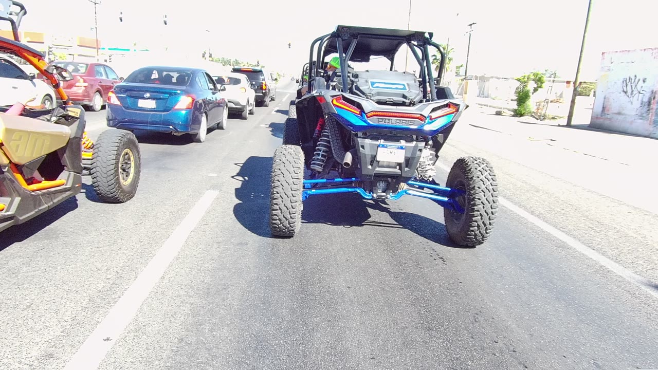 Blue and orange UTV centered in traffic on a paved road in Los Cabos, Mexico, with a blue car ahead, a yellow off-road vehicle at left, and no visible people.
