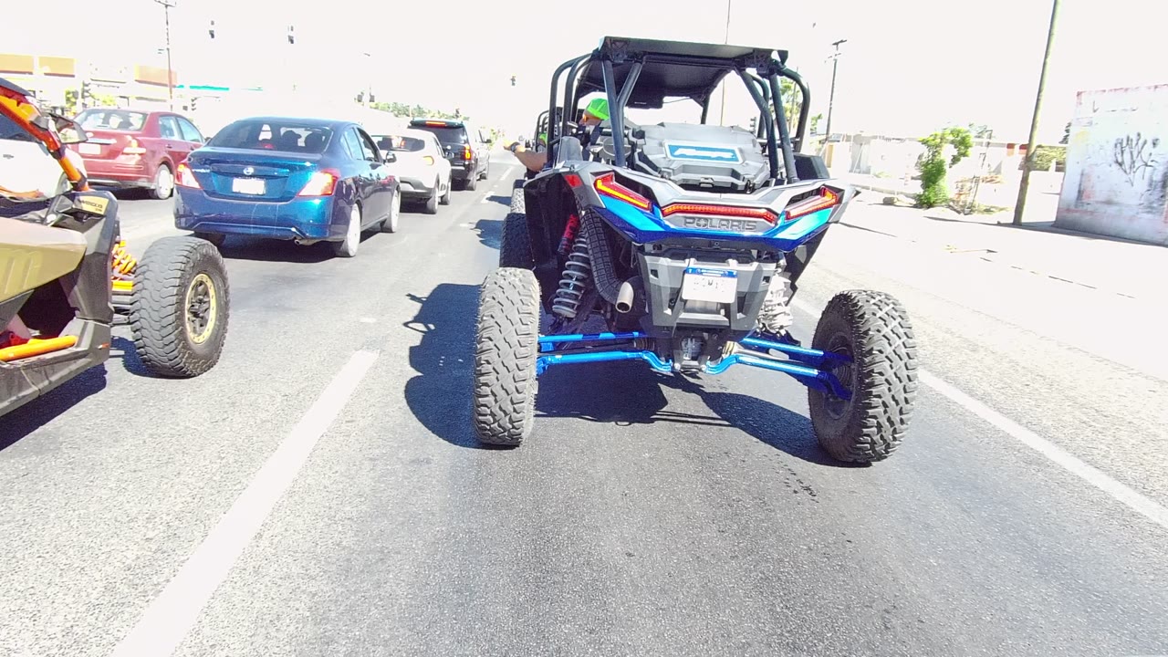 Rear view of the blue and orange UTV in the center of a paved Los Cabos traffic lane, with a blue car ahead and a bright wall and curb on the right, with no visible people.