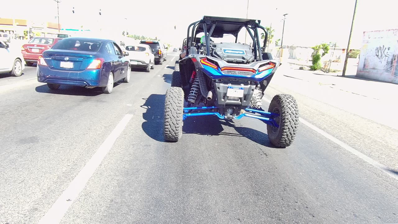 Blue UTV seen from behind in Los Cabos traffic with cars ahead, asphalt lane markings, bright roadside glare, and no visible people.