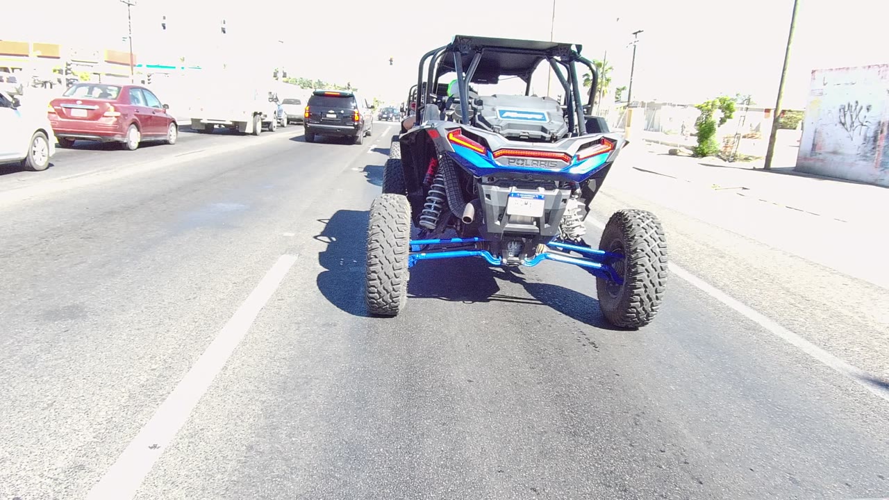 A blue UTV drives ahead on a sunlit Los Cabos road as red and black cars appear in adjacent lanes, with no visible people.