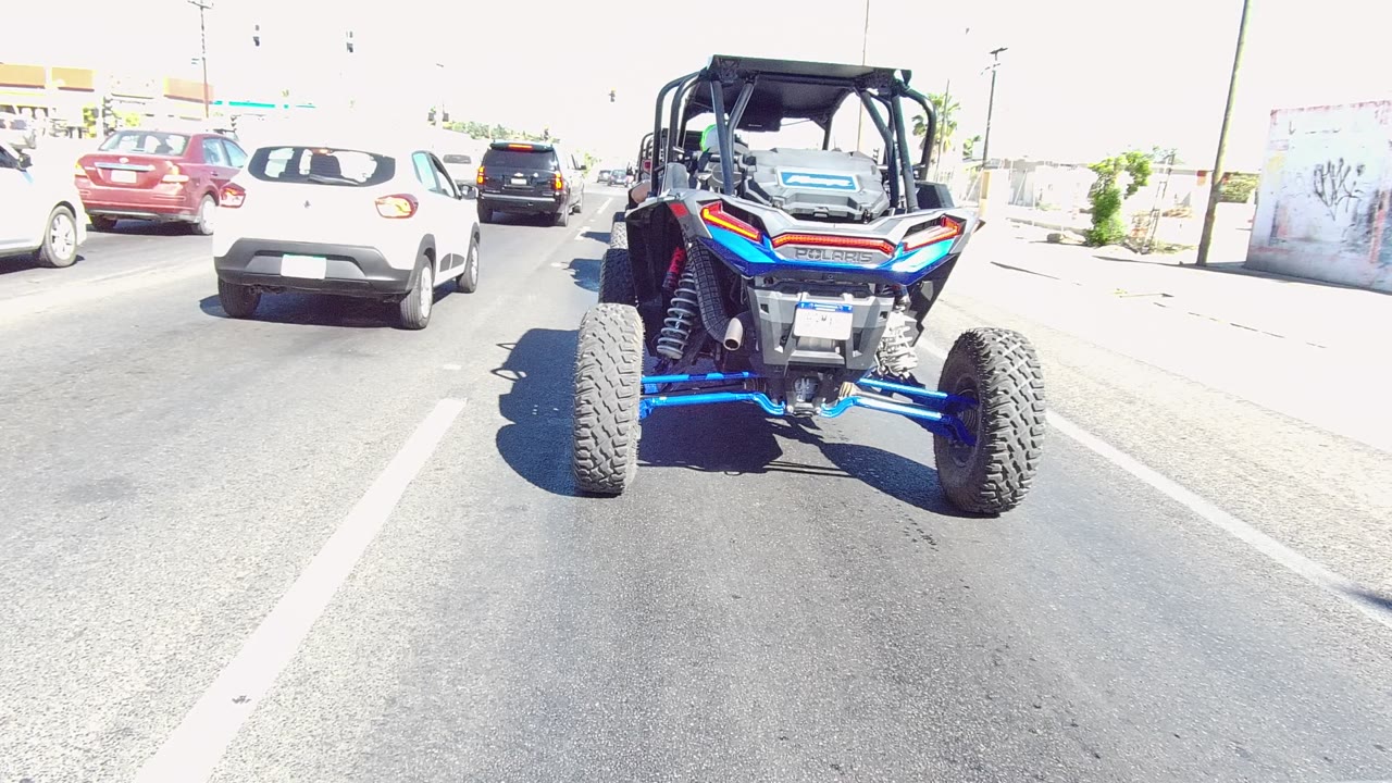 The blue UTV stays centered on a bright Los Cabos street while a white car moves farther ahead and other traffic fills the lane, with no visible people.