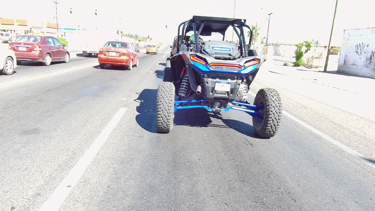 Rear view of a blue UTV on a bright Los Cabos road with red and white cars ahead and no visible people.