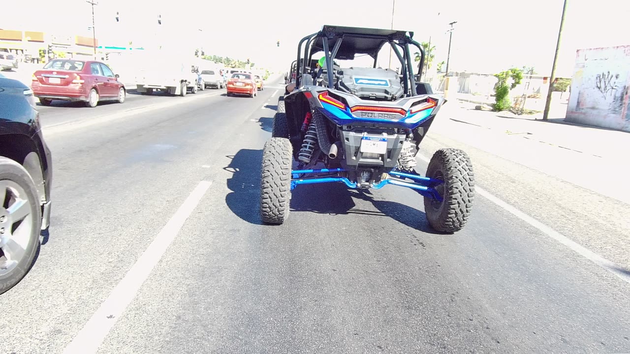 A black SUV moves into the right side of the frame beside the blue UTV on a Los Cabos road, with red cars ahead and no visible people.