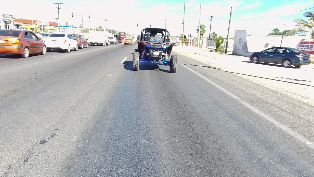 Blue UTV centered ahead on a Los Cabos urban road, with white and orange cars to the left, a dark car near the curb, bright pavement, and no visible people.
